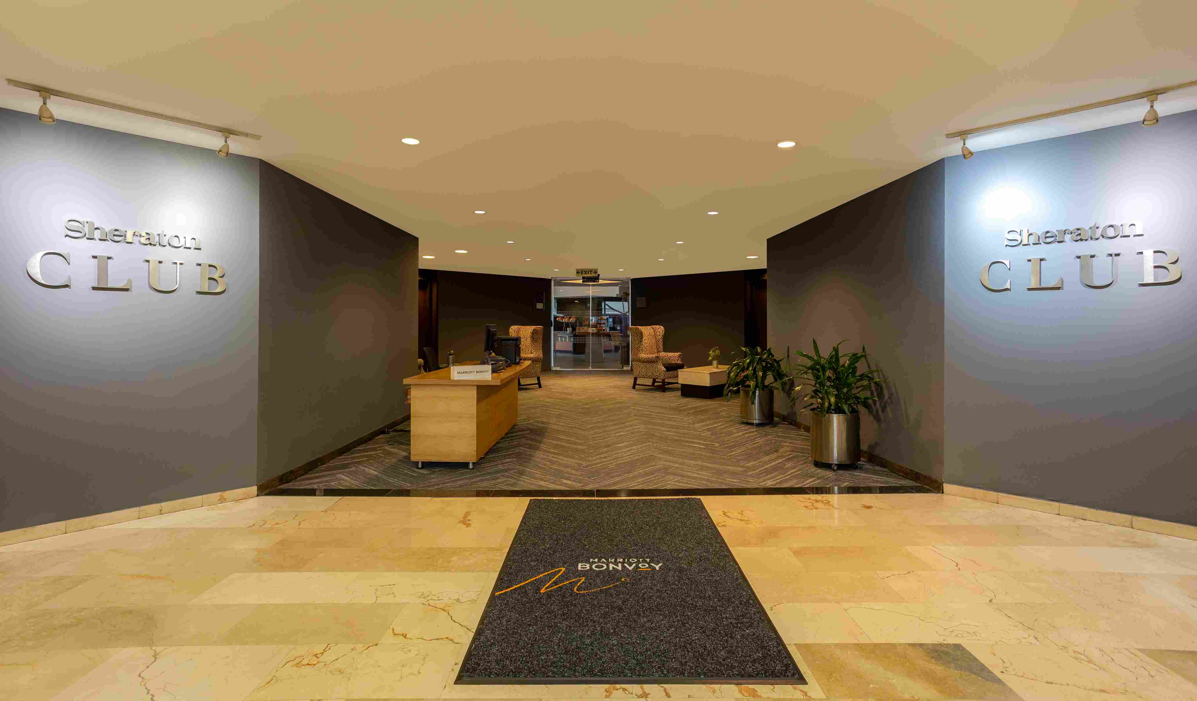 Corredor largo en el que hay una mesa de madera en la que hay un computador, dos plantas y dos poltronas. Hay dos paredes pintadas de gris en las que destacan leyendas que dicen: "Sheraton Club".