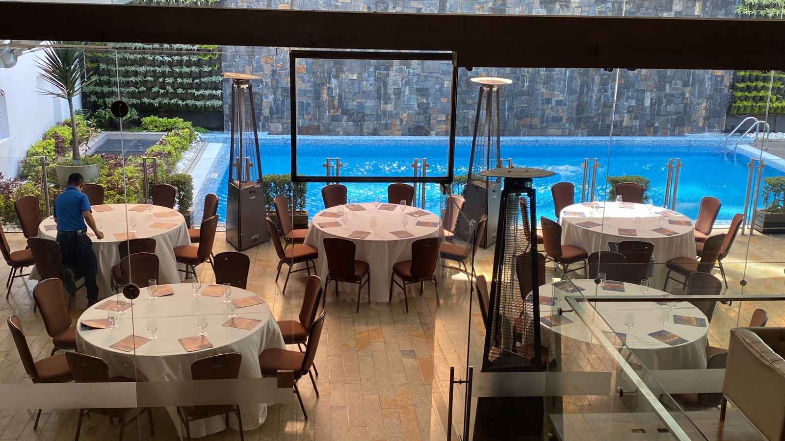 Salón con una piscina y al lado, separado por un vidrio un salón con mesas redondas dispuestas con manteles y copas de vidrio.