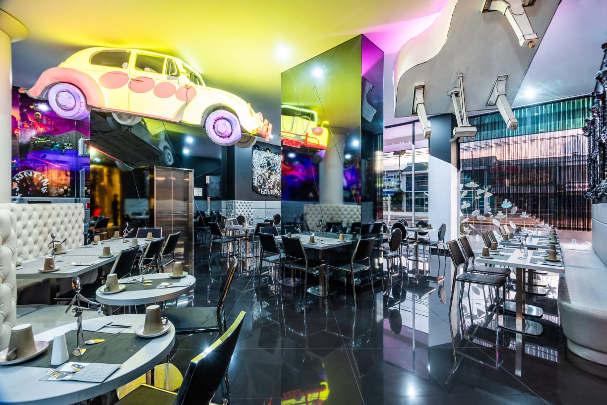 Interior de un salón para fiestas, decorado con un viejo Volkswagen y un piano blanco de cola que cuelgan del techo. Hay varias sillas metálicas y sofás que rodean pequeñas mesas circulares y cuadradas. Hay varios espejos en las paredes.