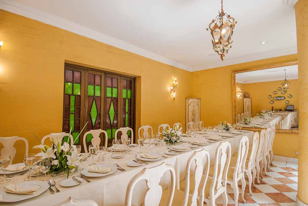  Salón con una larga mesa rectangular vestida con mantel color crema y varios platos y copas sobre ella. La mesa está rodeada por sillas de estilo antiguo.