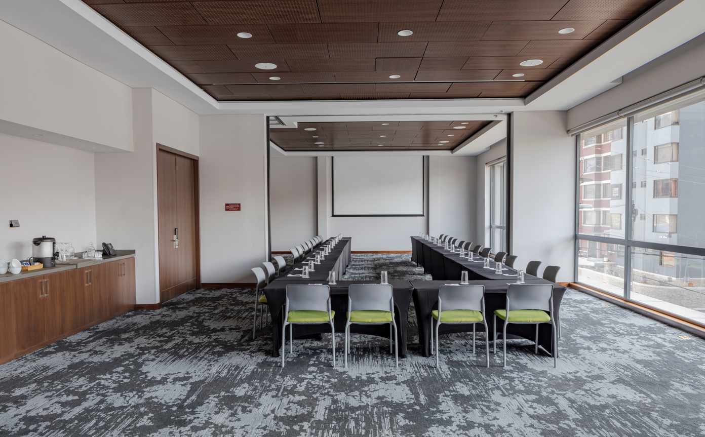 Salón mediano con varias mesas blancas formando una U. Hay varias sillas de color gris y amarillo alrededor. Al fondo hay una pantalla de viodeobeam y al lado una gran ventanal.