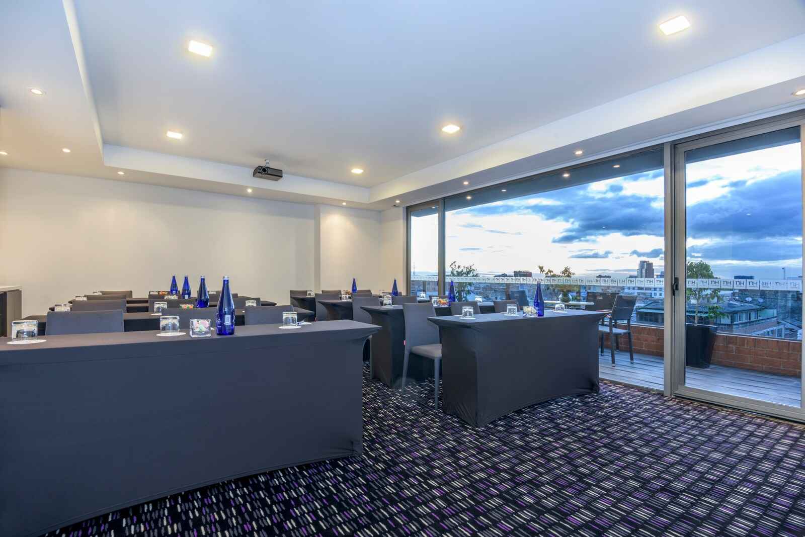  Un salón en un piso alto con varias mesas con mantel gris, botellas y vasos de vidrio. Hay una pantalla gigante y al fondo hay una ventana donde se observa la ciudad al atardecer. 