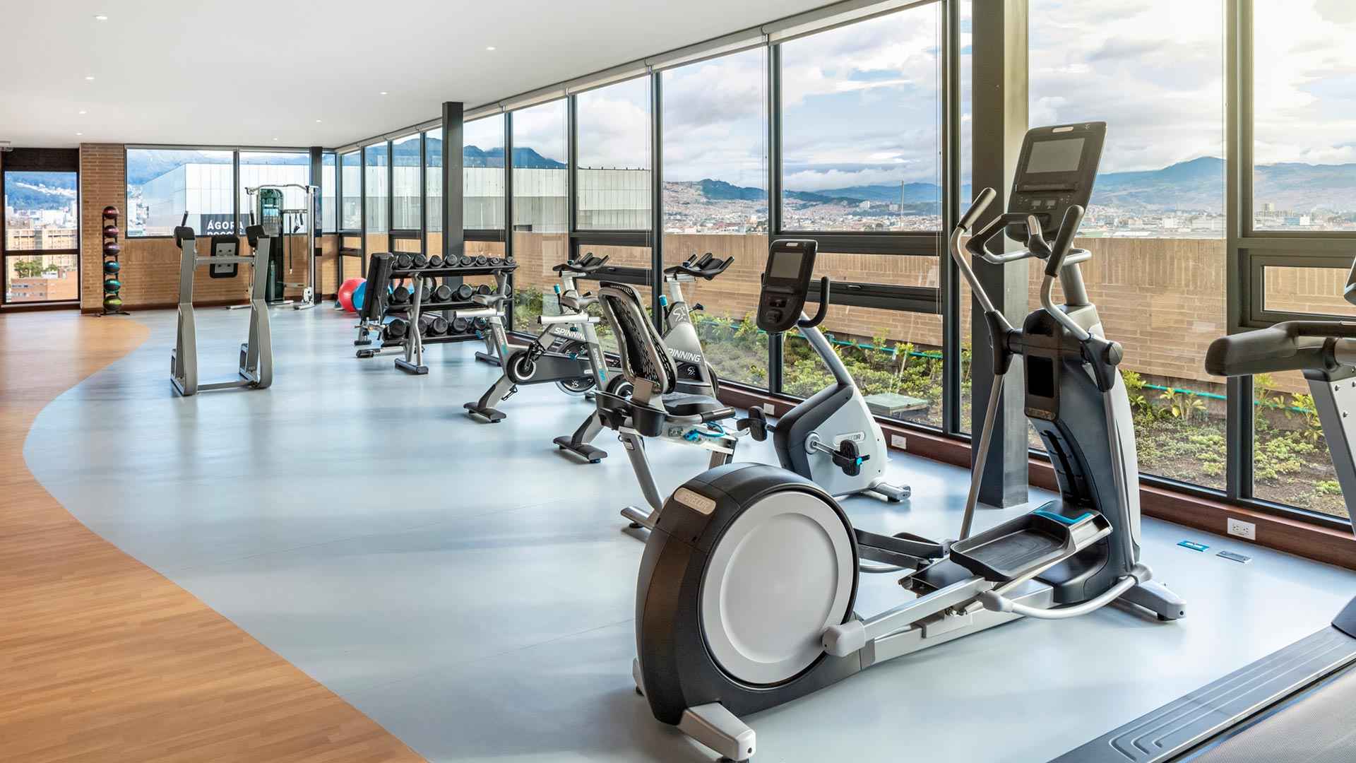 Sala de gimnasio cubierto, en un piso alto desde donde se ve la ciudad. Hay varias máquinas para hacer ejercicio.