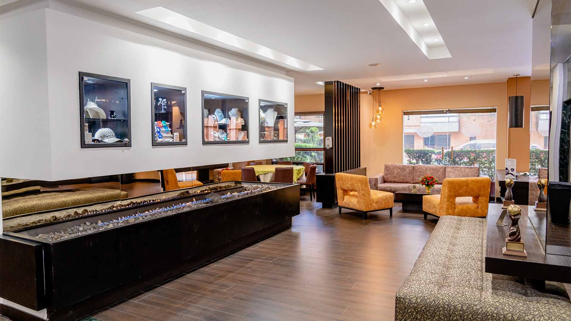 Un salón de recepción con varias poltronas, sofás y una gran chimenea rectangular. Encima de la chimenea hay una vitrina con varios suvenirs. Al fondo hay un ventanal hacia la calle.