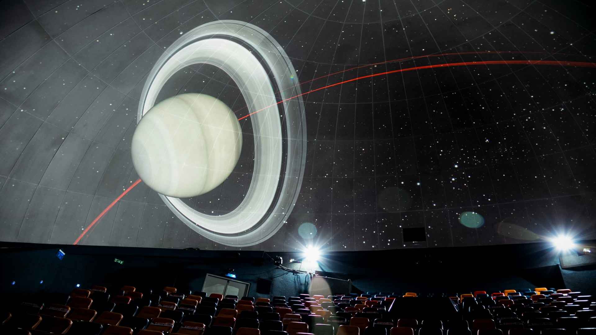 Interior de un salón que parece ser un cinema con una pantalla gigante, proyectando el planeta saturno y varias sillas al frente.