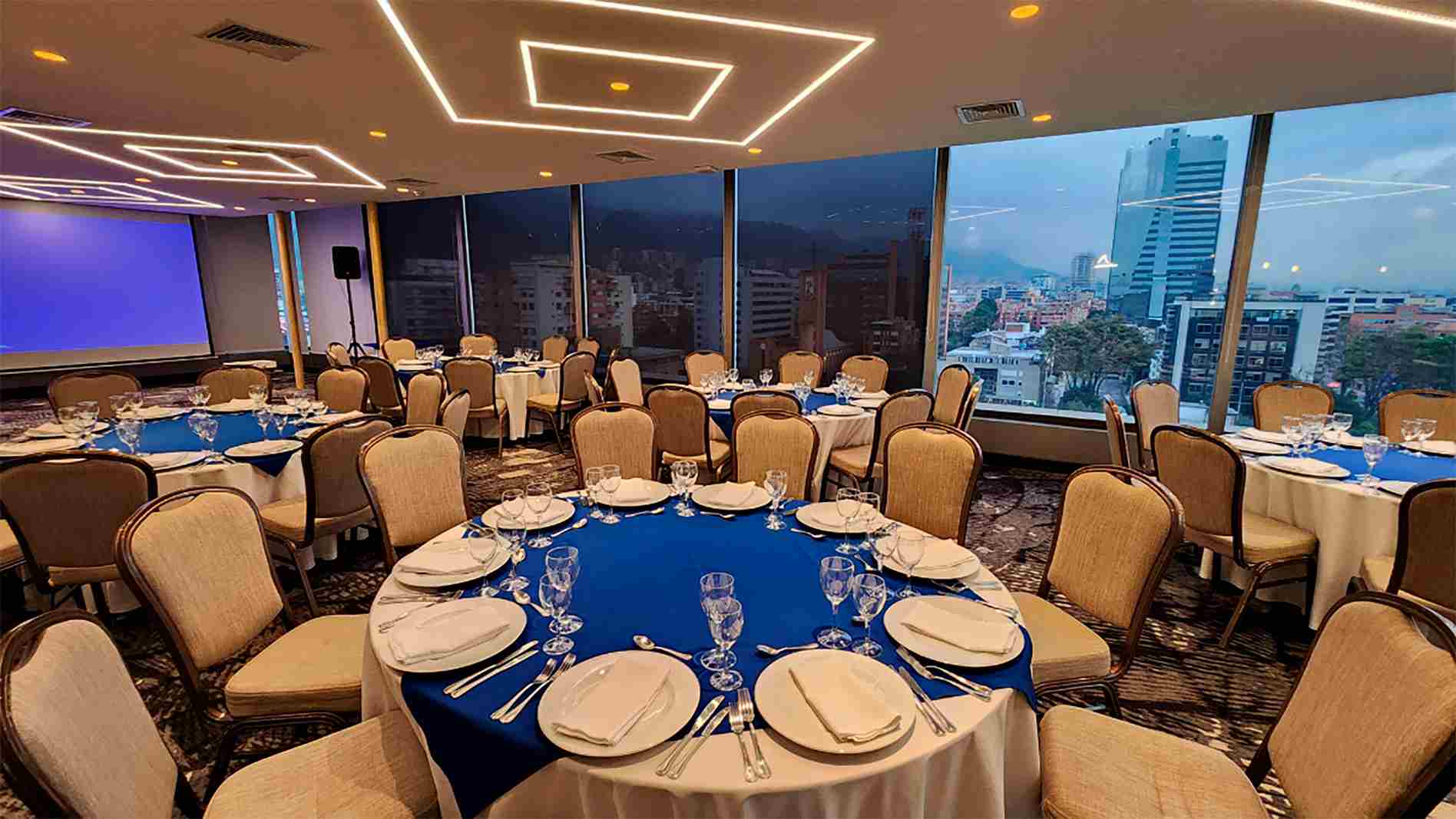 Salón en piso alto con mesas redondas y sillas alrededor. Las mesas están decoradas con manteles, cubiertos y copas de cristal. A un lado hay un gran ventanal y al fondo una pantalla de videobeam.