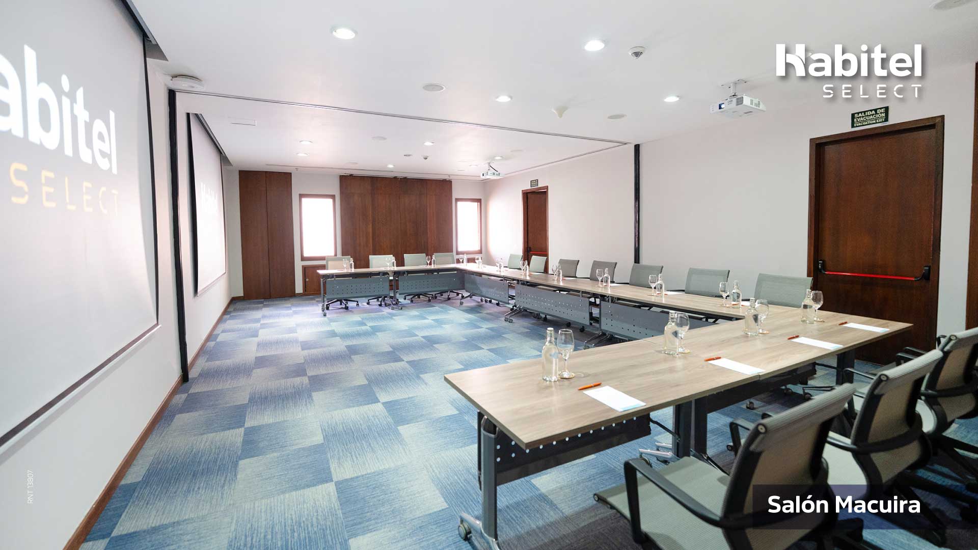 Salón con una gran mesa en forma de U. Alrededor de la mesa hay sillas y una pantalla de videobeam al frente. Al fondo hay dos ventanas.
