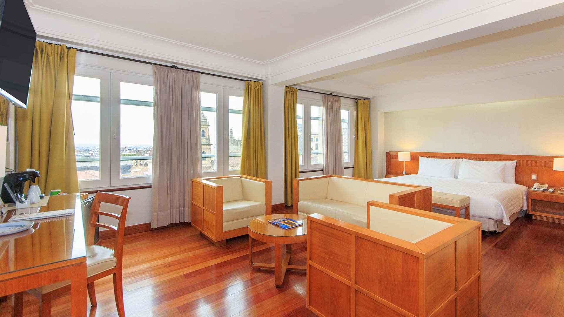 Habitación amplia con una cama grande y varias poltronas y piso de madera. Hay un televisor en una pared y un gran ventanal hacia la calle.