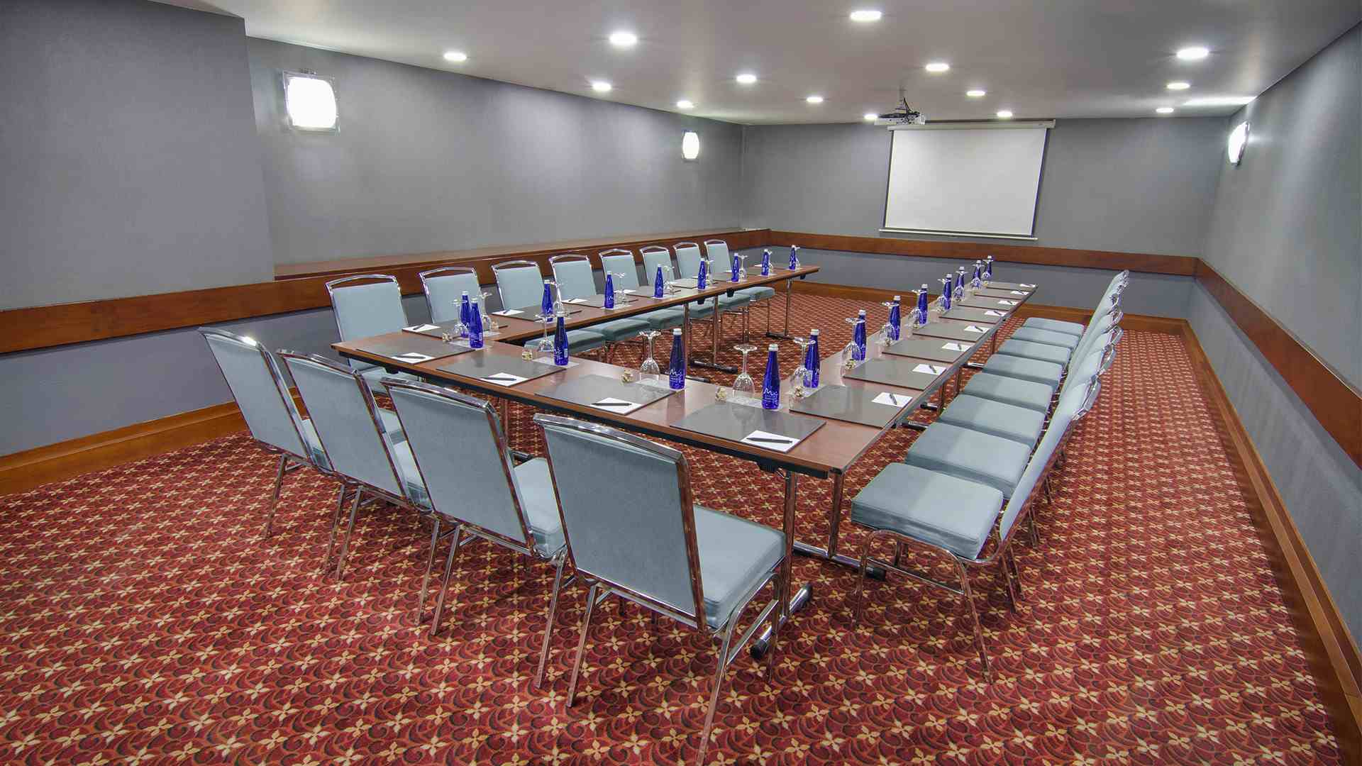 Salón rectangular con varias mesas formando una U. En las mesas hay botellas y copas de cristal, y están rodeadas por varias sillas metálicas.