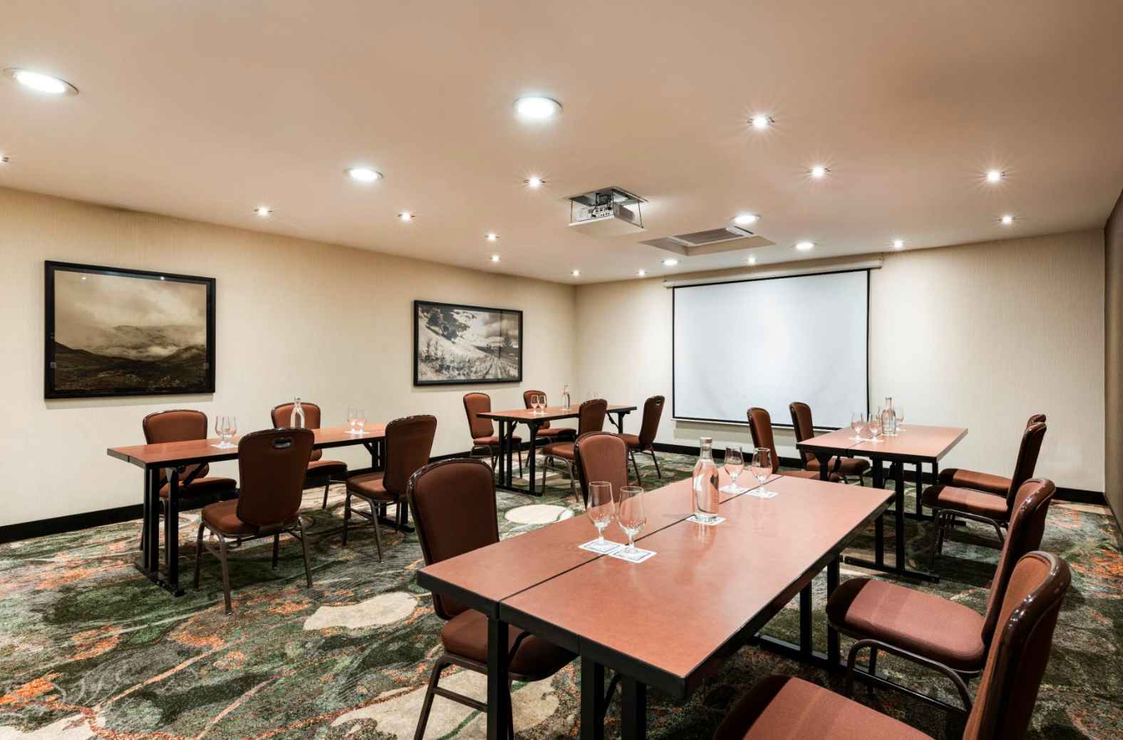 Salón con 4 mesas rectangulares con cuatro sillas cada una. En cada mesa hay cuatro copas y una botella de cristal. En las paredes hay cuadros y en una de ellas hay una pantalla de videobeam.