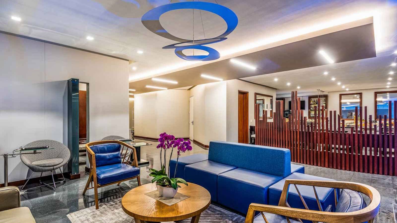 Salón como de recepción de un hotel donde hay un sillón grande, varias sillas y una mesa de centro con un arreglo floral.