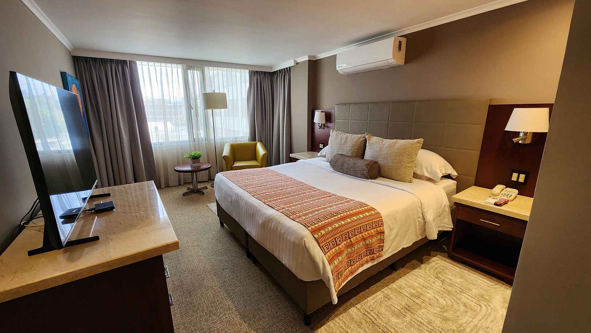  Cuarto de habitacion en el que hay una cama amplia, dos mesas de noche, una poltrona en cuero, una mesa redonda con una planta y una mesa con un televisor.