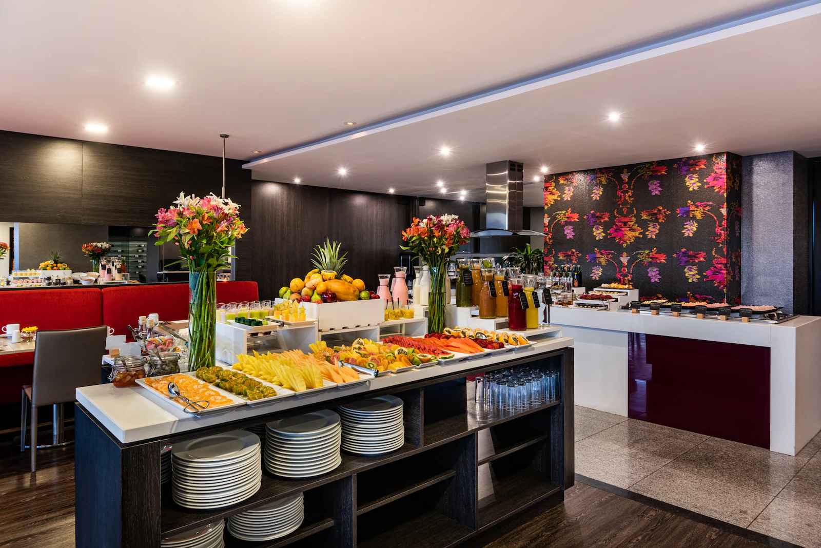 Interior de un salón con una barra repleta de pedazos de fruta, boetllas con jugo, platos, vasos y dos arreglos florales. 