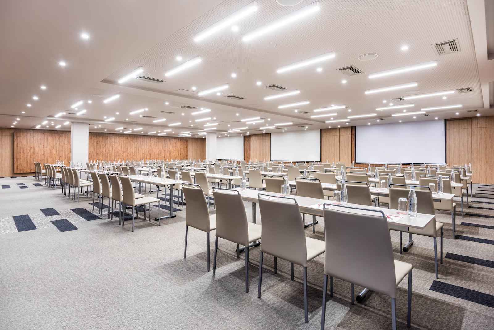  Salón amplio y entapetado dispuesto para conferencias con varias mesas y sus sillas organizadas de manera horizontal mirando al frente hacia una pantalla de proyector.