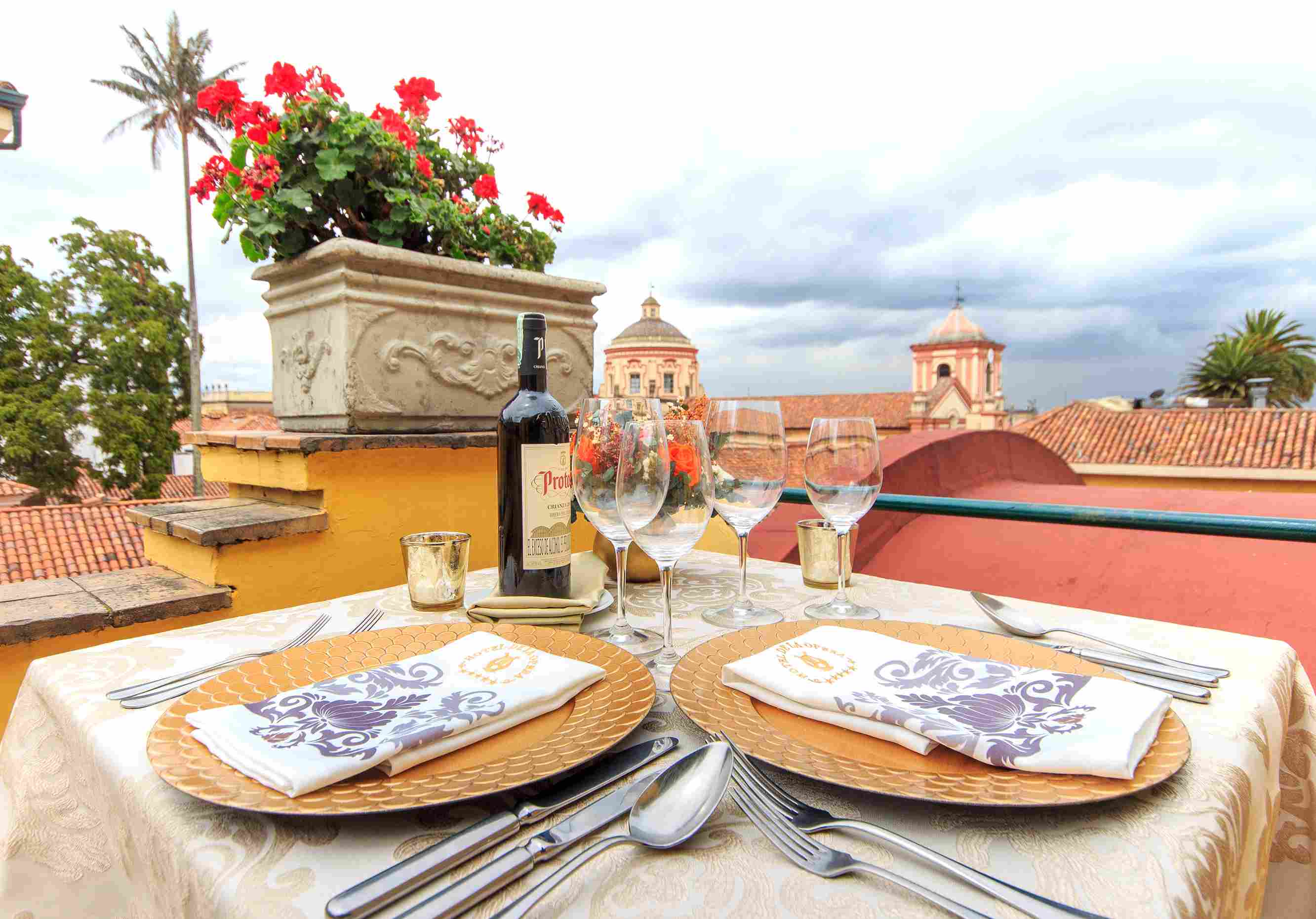  Mesa con dos platos, varias copas y una botella en una terraza exterior. Se observan techos y cúpulas de una iglesia cercana.
