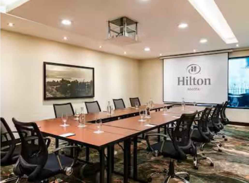 Salón con mesas organizadas en forma de U. Hay varias sillas rodeando la mesa y en ella varias copas de cristal. Al frente hay una pantalla de videobeam con una leyenda que dice: "Hilton".