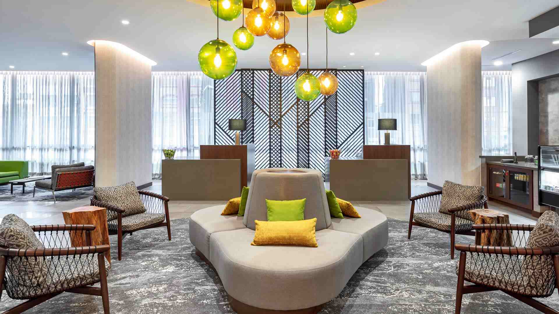 Salón como un lobby elegante con cuatro sillas de madera y cuero, dos troncos de madera y un sofá grande gris en forma ovalada y con cojines de colores. Encima hay una lámpara con globos de colores. A un lado se ve una vitrina con algunos suvenirs y al fondo un ventanal grande por donde ingresa luz natural.