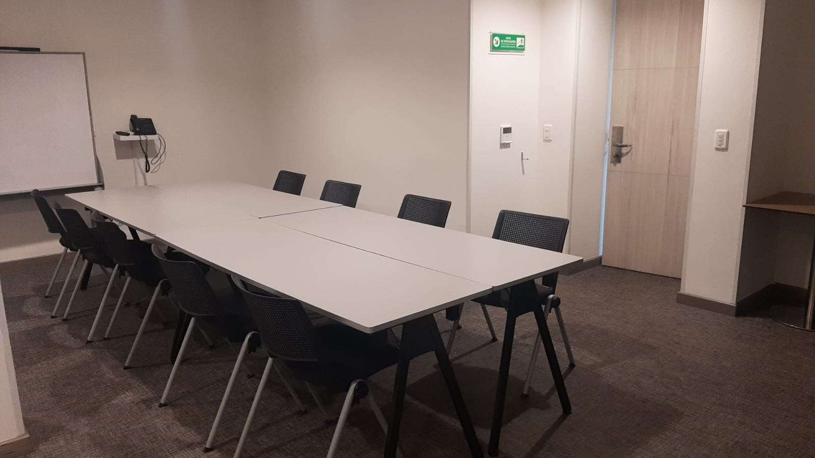  Pequeño salón con una mesa rectangular rodeada por 8 sillas, un teléfono fijo y una pantalla de videobeam.