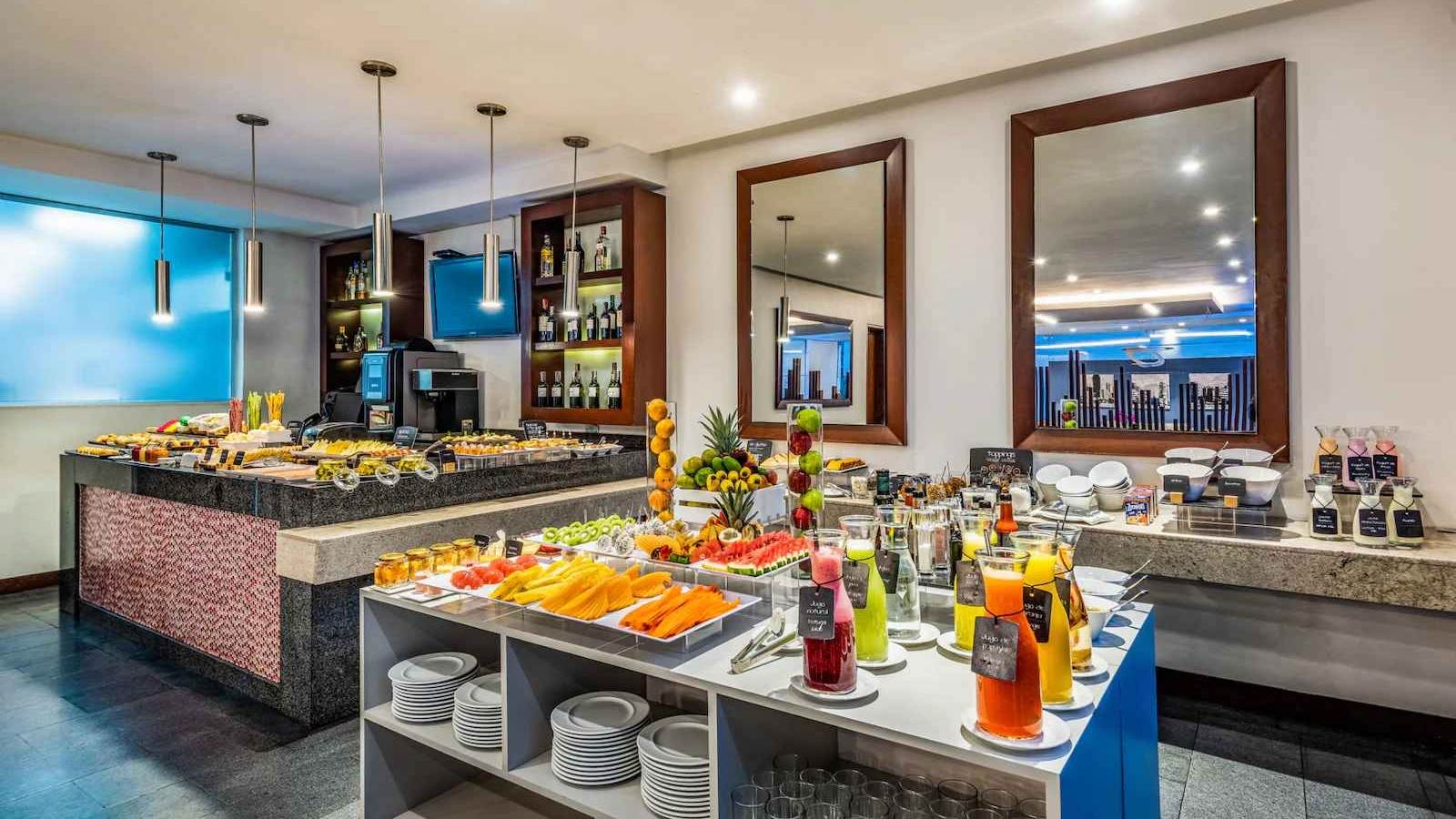  Salón de restaurante donde hay una mesa principal repleta de trozos de fruta, botellas con jugo, panes, platos y cubiertos. 