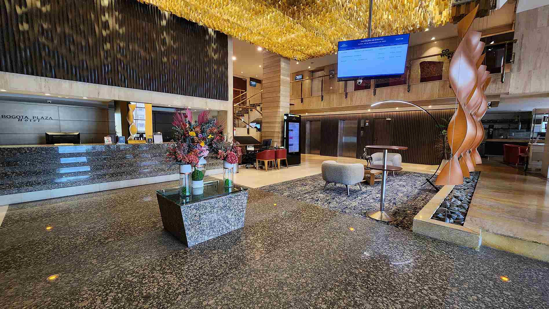 Salón estilo recepción en el que destaca el piso en marmol gris y brillante, una mesa en marmol con arreglos florales y un cubículo para atender. Hay una figura geométrica que cuelga del techo y una pantalla de televisión.