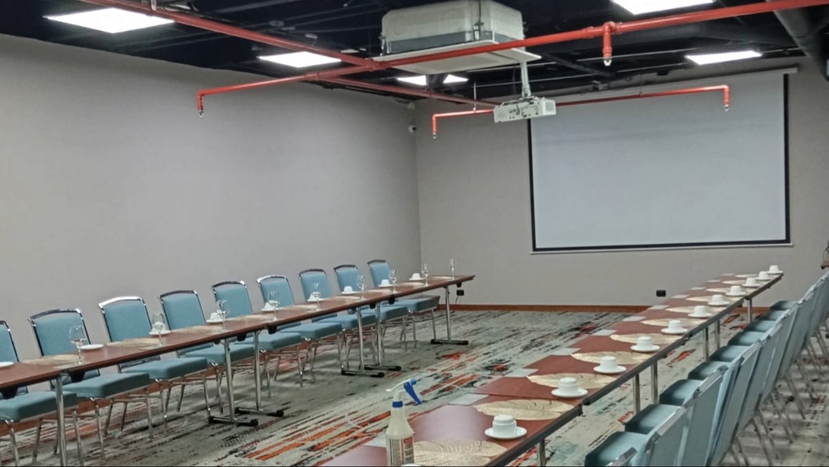 Amplio salón con dos filas de mesas y sillas una enfrente de la otra. En las mesas hay tasas y copas de cristal. Al frente hay una pantalla de videobeam.