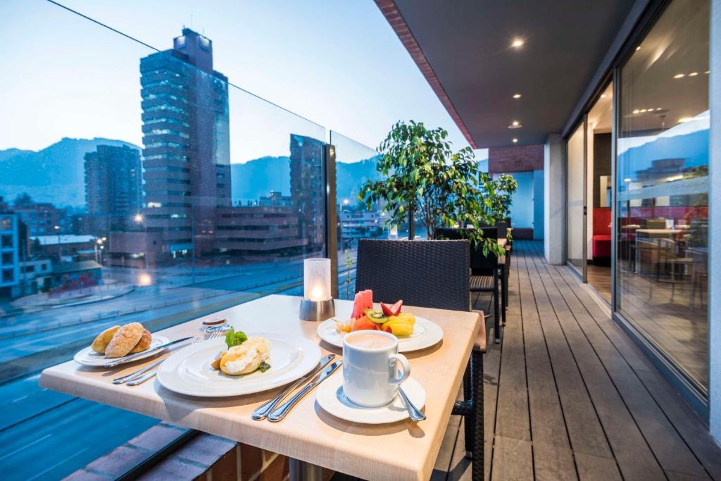 Terraza exterior en un piso alto en el que se observa una mesa con unos platos con fruta y panes, y un pocillo con un líquido que parece café. 