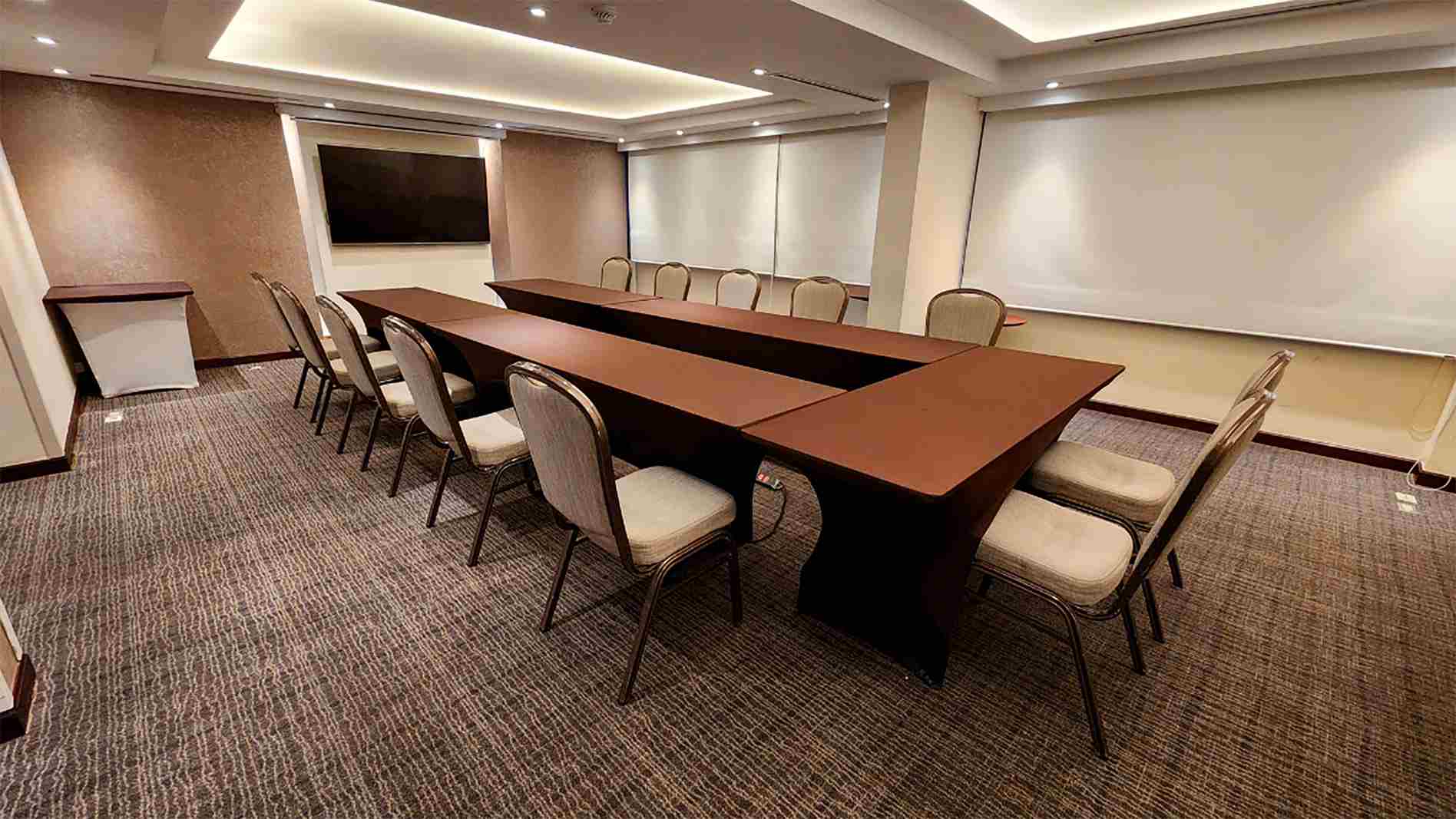 Salon con una gran mesa en U, decorada con manteles de color marrón. Alrededor de la mesa hay varias sillas blancas. Al frente hay una pantalla de televisión.