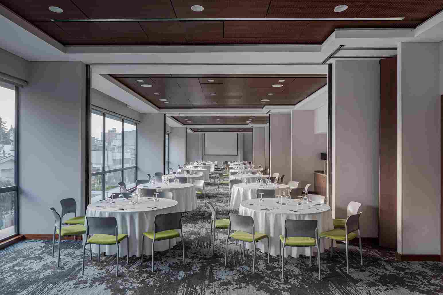  Gran salon rectangular con varias mesas redondas decoradas con manteles blancos y varias copas y cubiertos encima. Hay muchas ventanas a un costado. Al fondo hay una pantalla de viodeobeam.