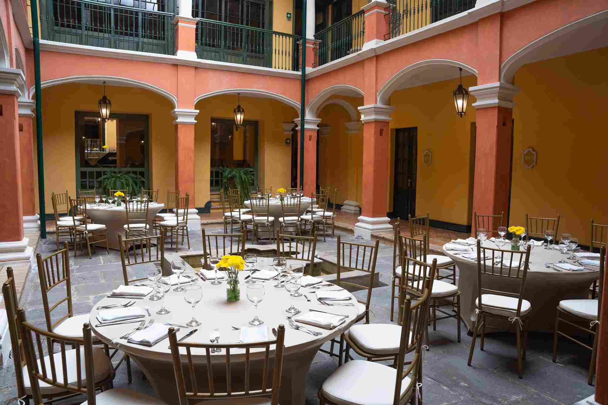  Patio interior de un recinto antiguo con paredes en arcos, donde hay varias mesas redondas, rodeadas de sillas de madera. En las mesas hay copas, platos y cubiertos.