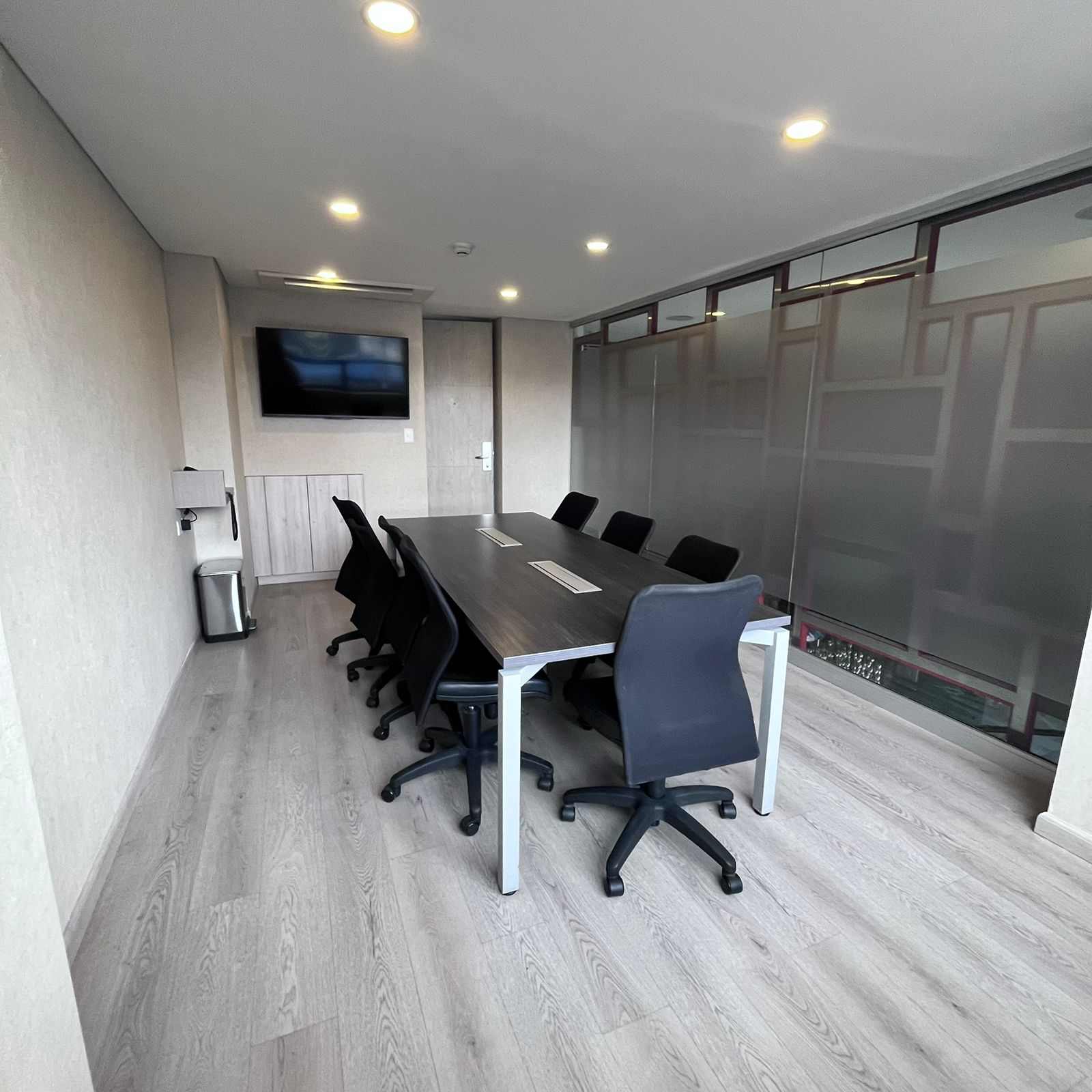 Pequeño salón con una mesa rectangular rodeada por 7 sillas, un y una pantalla de televisión.