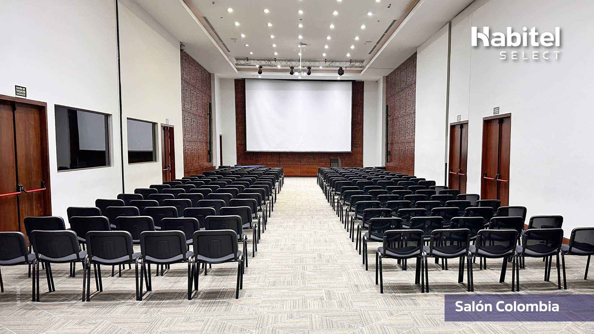 Salón rectangular en el que hay por lo menos un centenar de sillas, todas mirando hacia una pantalla de videobeam. 
