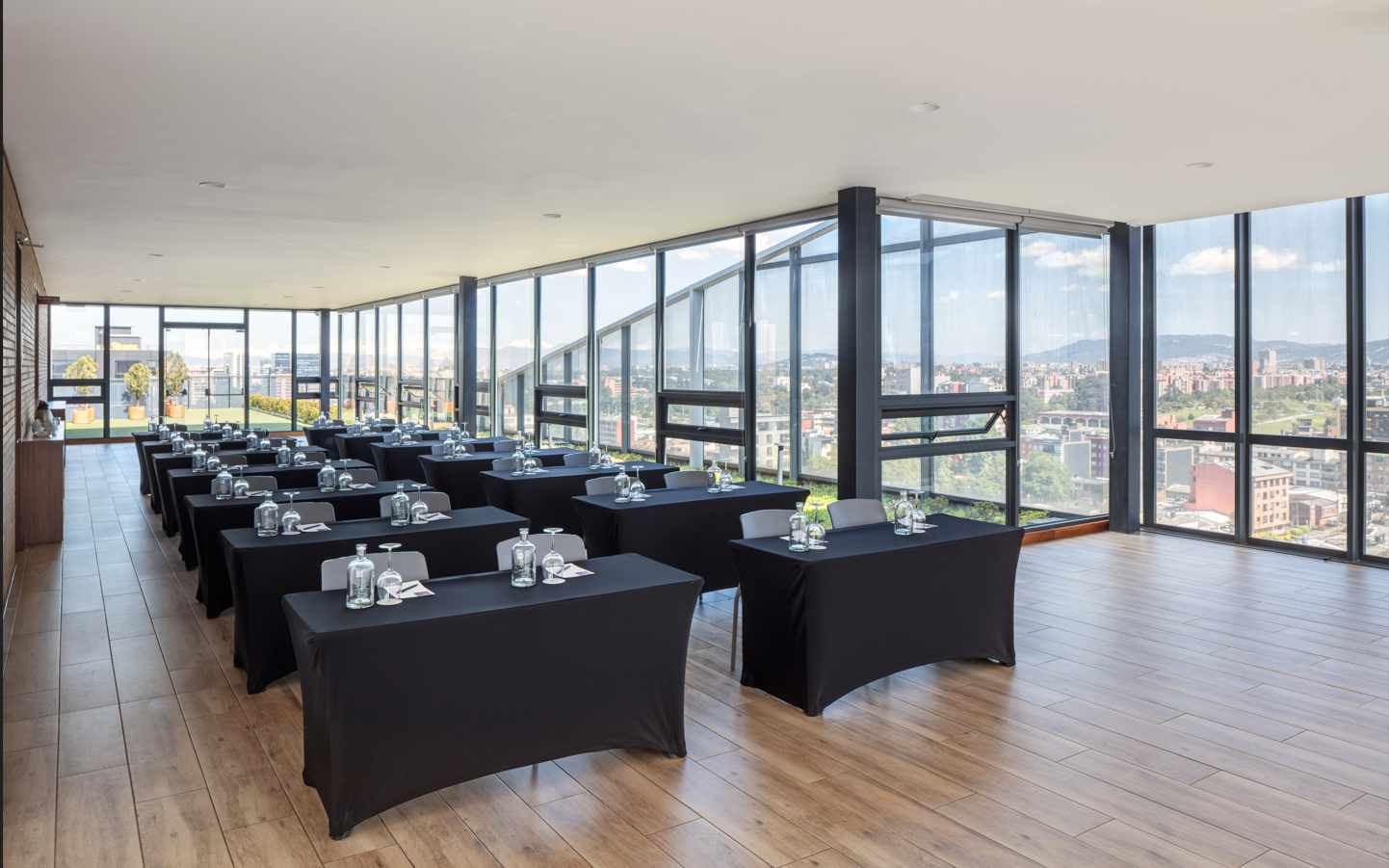  En una terraza hay casi veinte mesas decoradas con mantel negro. En ellas hay copas y botellas de cristal. por entre unos grandes ventanales se observa a lo lejos una ciudad.
