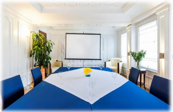  Pequeño salón con una gran mesa vestida con un mantel azul. Hay dos poltronas y aun lado un ventanal, parece que hacia la calle.