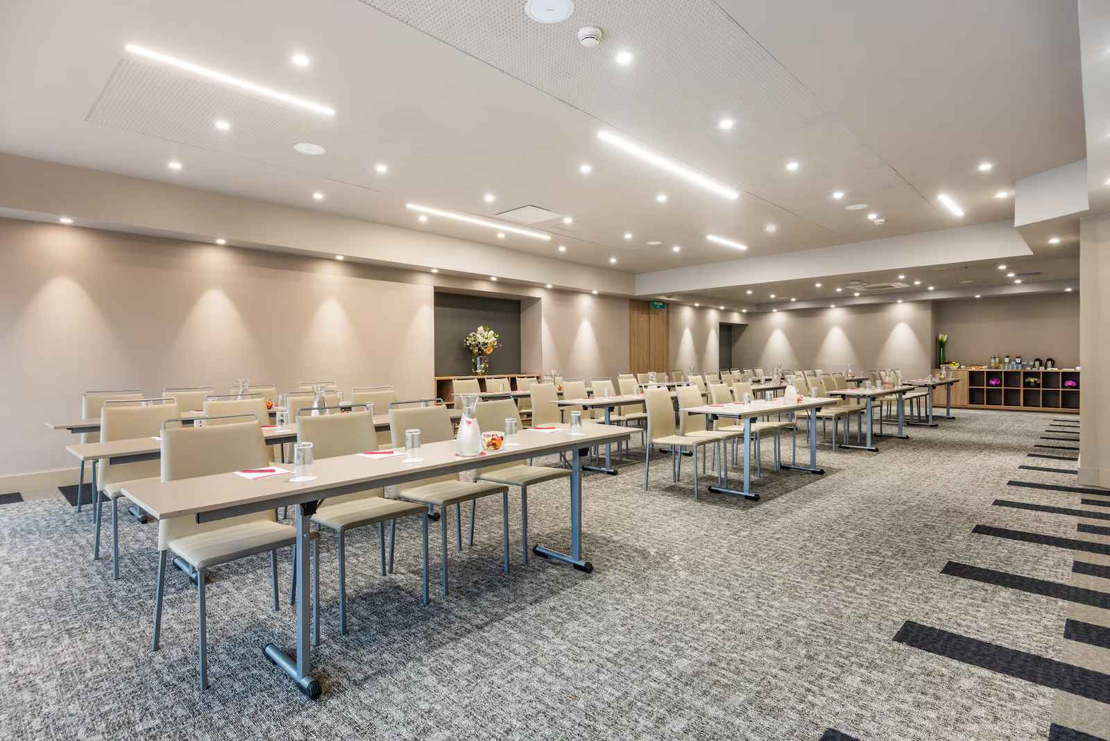 Salón entapetado con tres filas de mesas organizadas de manera horizontal. En las mesas hay vasos.