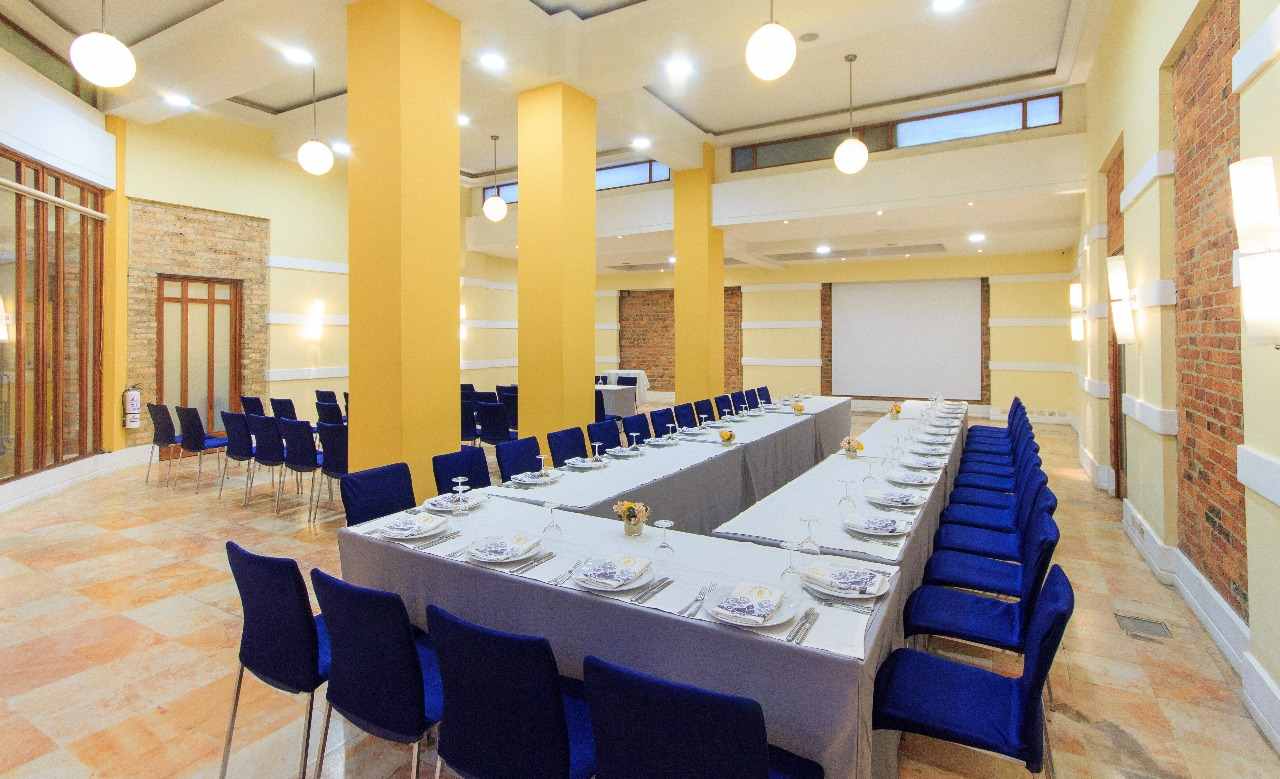 Gran salón con una larga mesa en forma de U y vestida con mantel blanco. Alrededor hay sillas vestidas con terciopelo azul. Sobre la mesa hay platos y copas.