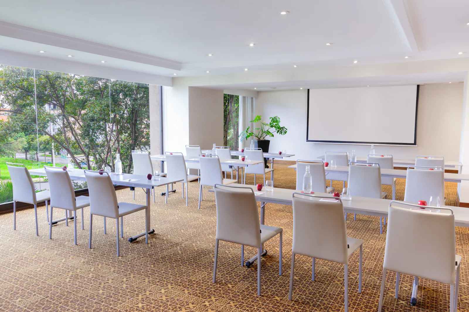Salón con ventanales a jardines a los lados. Hay dos filas con 6 mesas. En cada mesa hay manzanas y botellas de agua. Al frente hay una pantalla de videobeam y una matera con una planta.