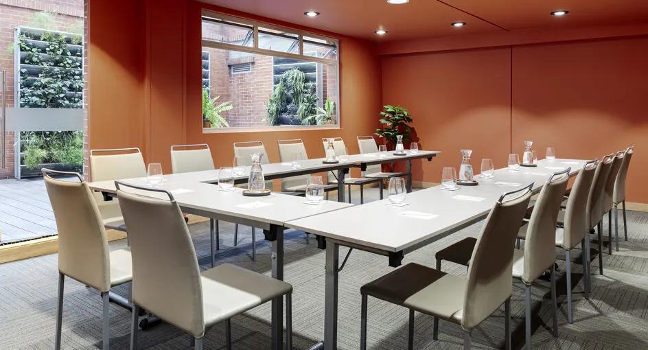Salón con mesas blancas organizadas en forma de U y rodeadas por varias sillas también blancas. El salón tiene las paredes pintadas de naranja y cuenta con una ventana y una puerta en vidrio.