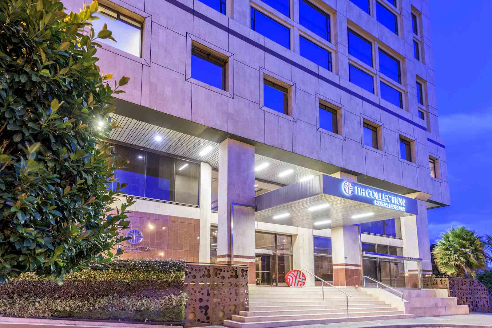  Fachada de edificio en el que sobresale una fuerte luz azul de atardecer. El edificio tiene muchas ventanas. En la parte baja de la pared principal hay un aviso en letras de neón que dice: "nH Collection"