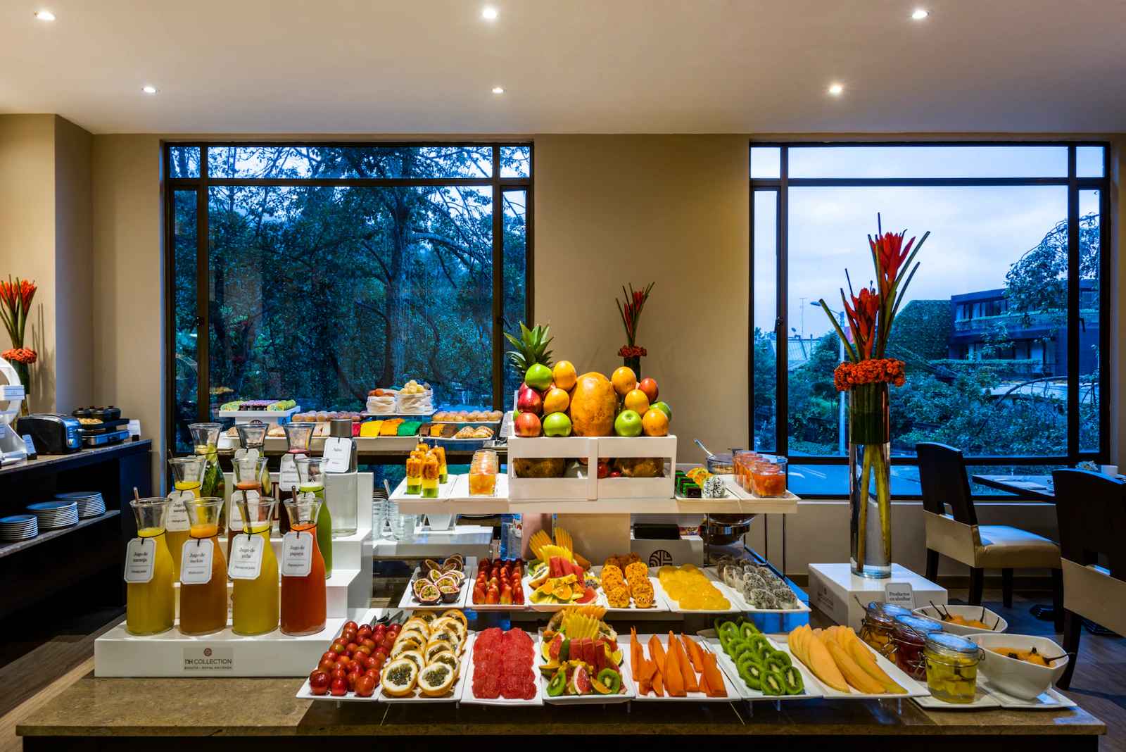  Salón en el que hay una mesa amplia sobre la que hay trozos de frutas y botellas de jugo. Al fondo hay dos ventanales hacia la calle.
