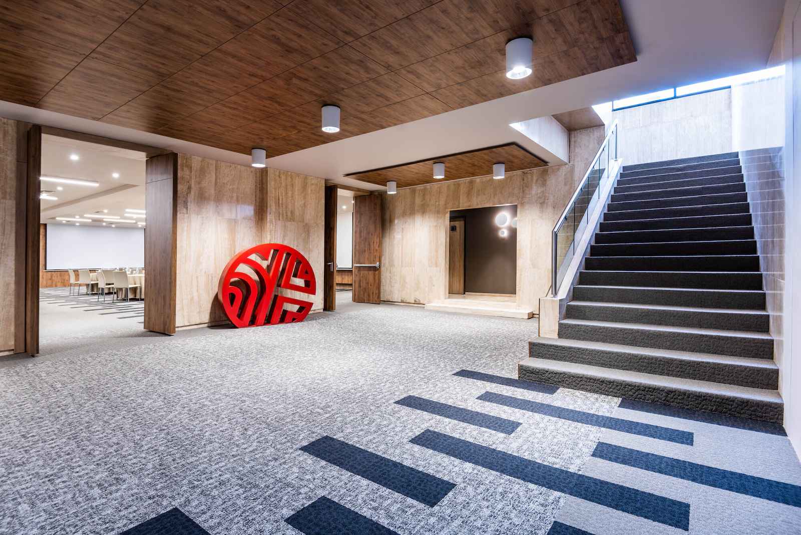 Salón como un corredor donde hay una escalera y una entrada a otro salón.  El piso tiene tapete y el techo está cubierto con madera.