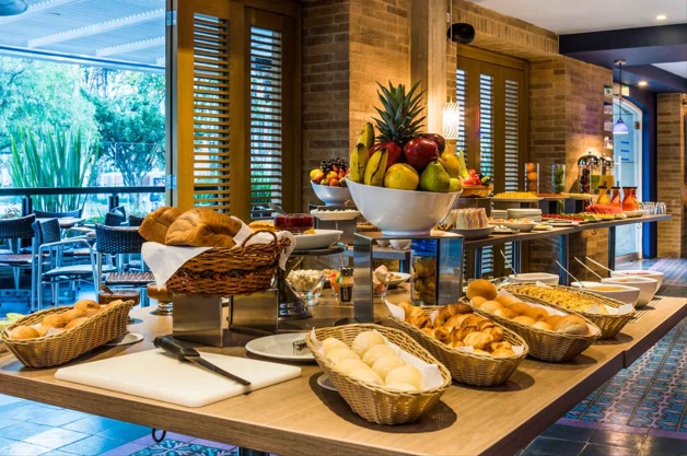 Una mesa amplia en un salón que parece un comedor, sobre la que hay trozos de pan, frutas y botellas de jugo. Se observa a un lado una vista hacia una terraza exterior en un jardín.