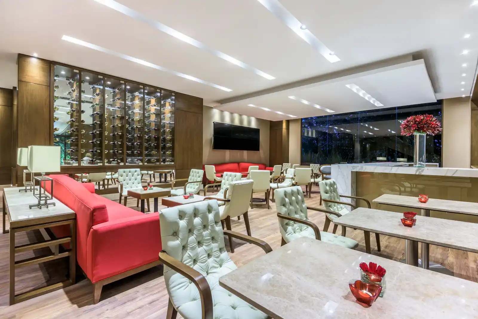 Salón en el que sobresalen varios sofás color rosa, sillas blancas abullonadas. Hay una pantalla de televisión en una pared y una gran cava de cristal y madera con varias botellas.