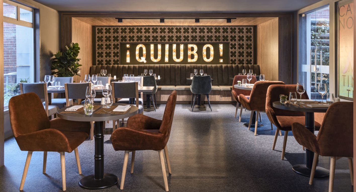  Salón en el que destaca un gran aviso en neón con una leyenda que dice: "¡Quiubo!". Hay varias mesas y sillas forradas en terciopelo café.