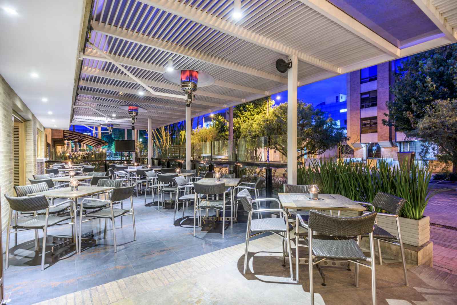 Terraza exterior en primer piso con varias mesas y sillas alrededor. Hay dos calefactores y una pantalla de televisión.