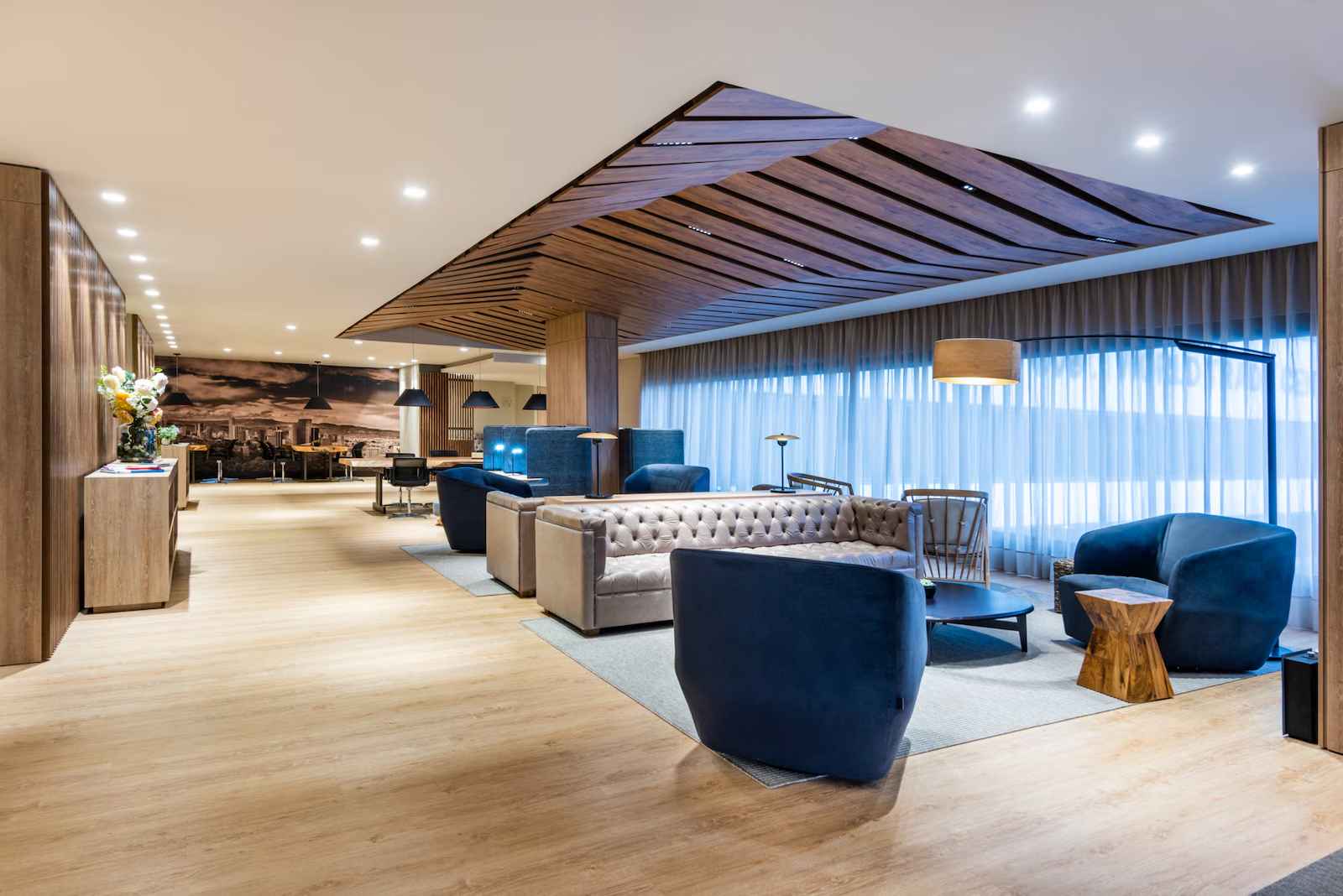 Salón estilo recepción donde hay varias poltronas, sofás y lámparas. Techo y paredes tienen decoraciones de madera.