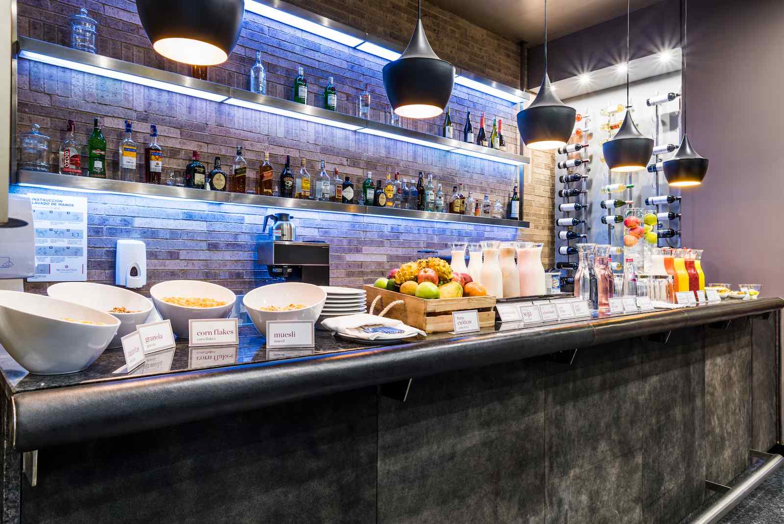 Salón pequeño con un mesón con varios pedazos de fruta, cereales y varias botellas con jugo. Al fondo en una pared hay una estantería con varias botellas de licor.