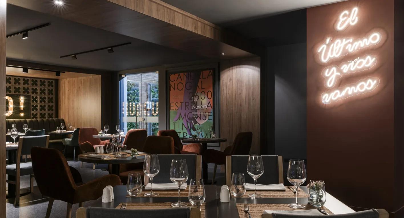  Salón en el que destaca un gran aviso en neón con una leyenda que dice: "El último y nos vamos". Hay varias mesas con copas y vasos de cristal, y sillas forradas en terciopelo café.