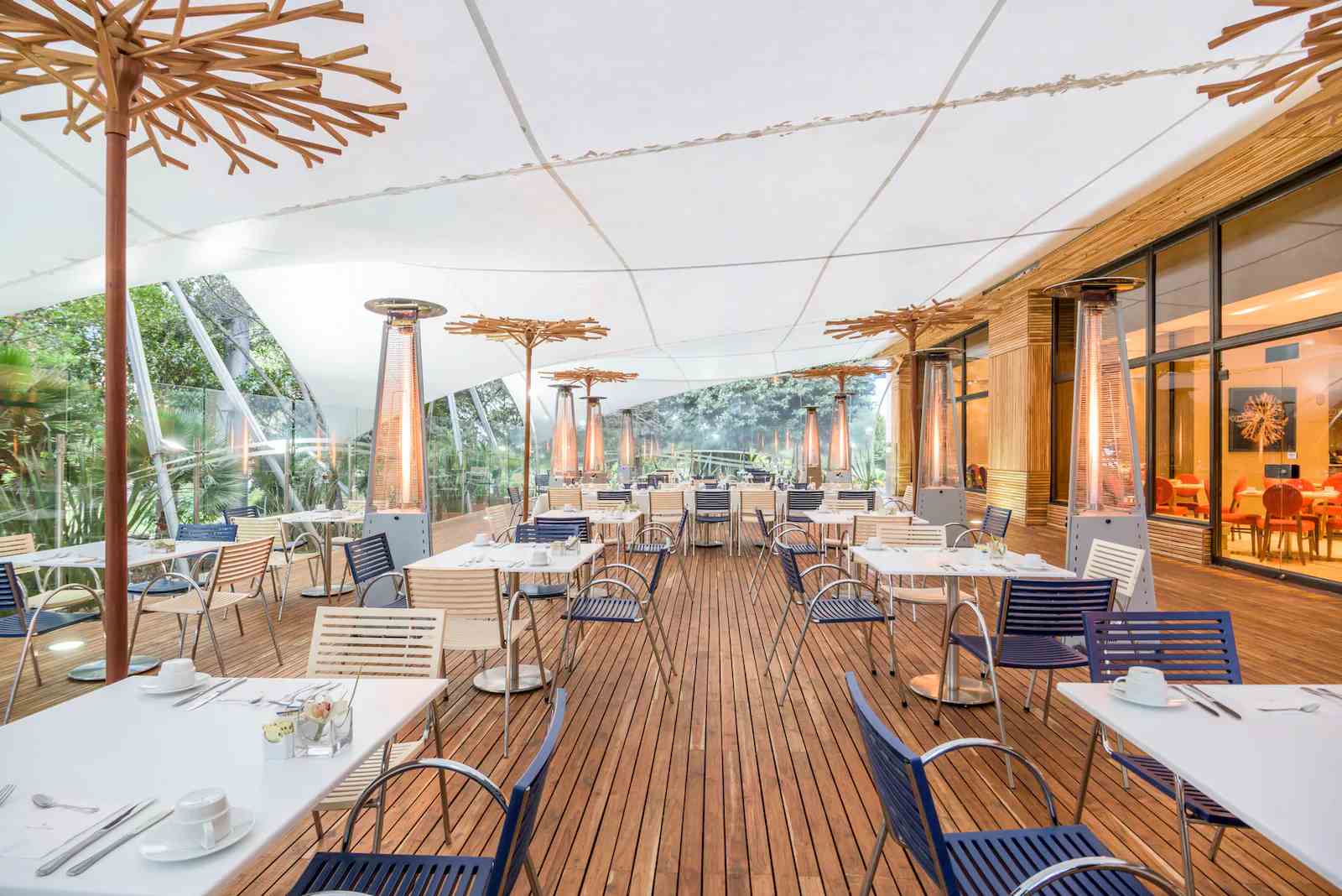 Terraza interior decorada con piso en madera y un techo en lona blanca. Hay varias mesas con sillas rodeadas por varios calentadores de ambiente en forma de triángulo.