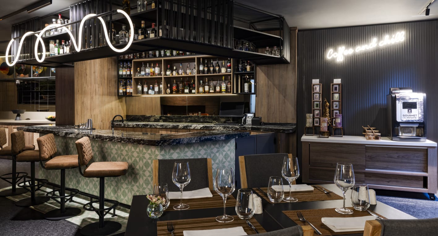 Salón tipo bar en el que sobresalen una barra y una estantería con varias botellas de licor.  Hay una mesas cuadrada con copas y vasos de cristal. Al lado de la barra hay cuatro sillas de cuero.