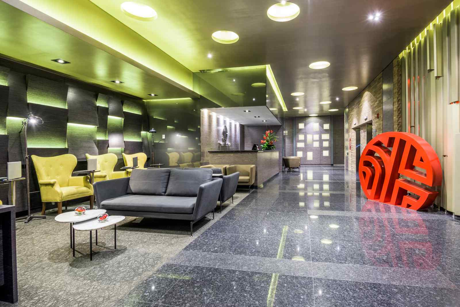 Salón largo rectangular, estilo recepción en el que sobresalen varios sofás grises y poltronas amarillas. El piso es de cerámica brillante.